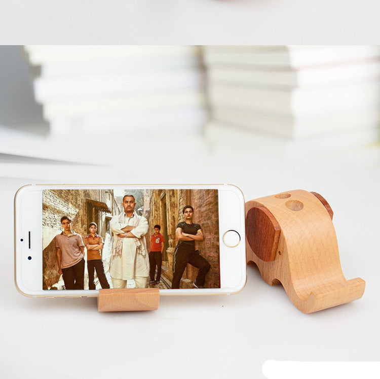 Lifestyle scene showing wooden elephant phone stand holding a smartphone on a desk with books
